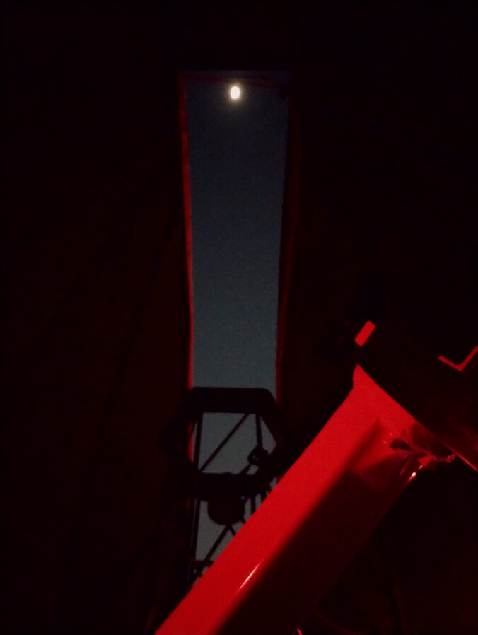 Silhouette of a telescope in red light image of the moon through the observatory doom slit