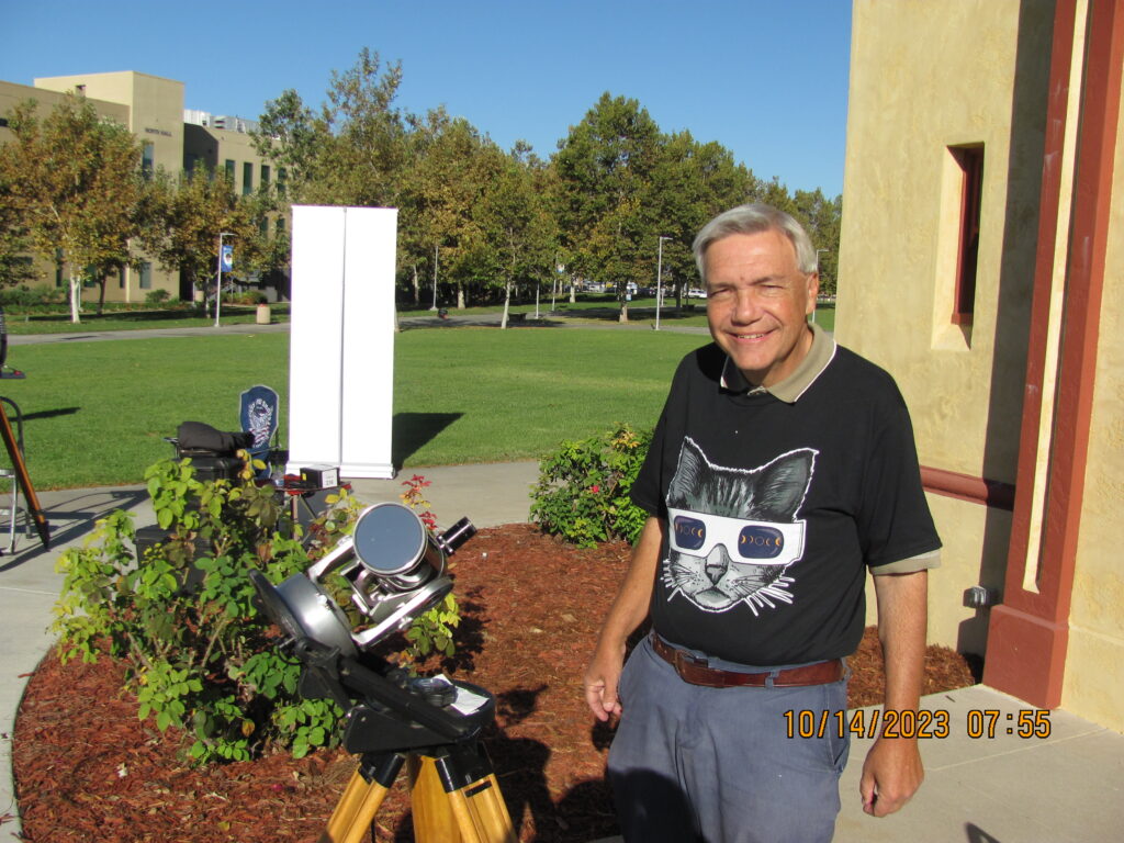 man looking at the camera with a cat wearing solar filter eyeglasses image on a t-shirt with a telescope to his right