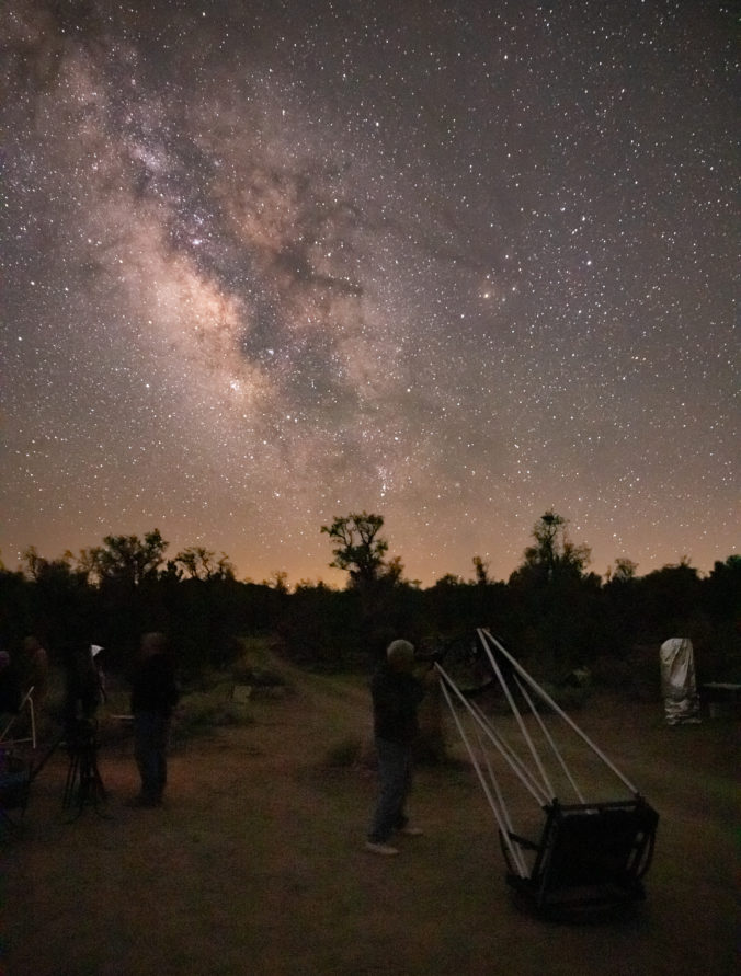 A man looking through a telescope pointed to the Milky Way galaxy in the sky with trees on the horizon