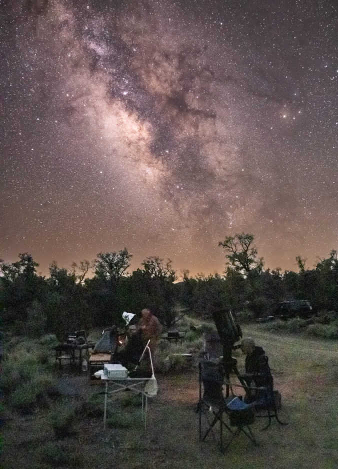 Two men at telescopes with the Milky Way galaxy in the sky with trees on the horizon