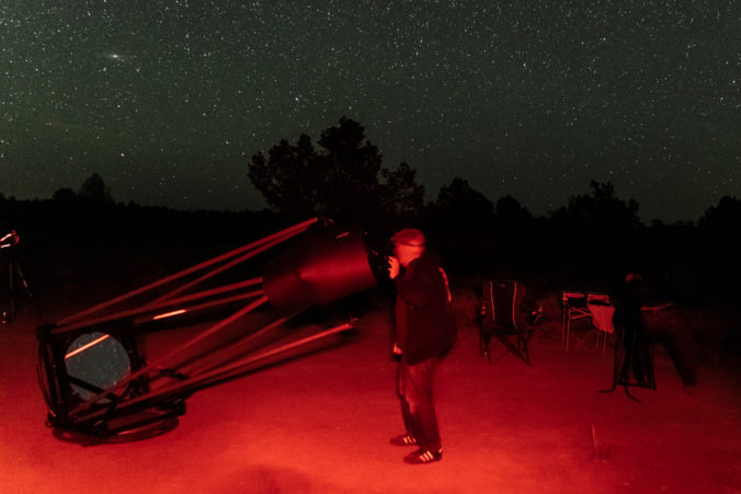 A man viewing through a telescope with the ground lite in red light. The stars and Andromeda galaxy visible in the sky and trees on the horizon