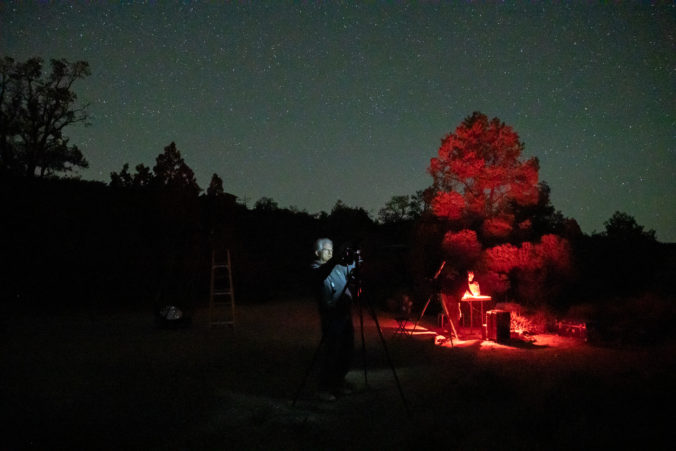 Two men illuminated, one in red light, in the dark with stars in the sky and trees in the background