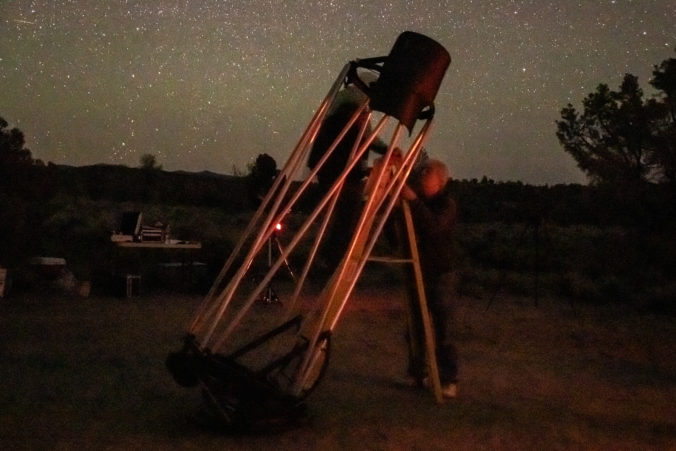 A man climbing a latter viewing through a telescope while another holds the latter. Stars in the sky with trees in the background
