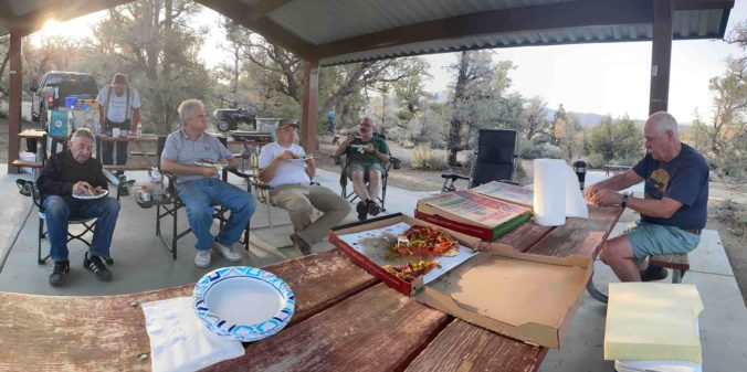 Six men sitting around picnic table eating pizza under an awning with trees in the background